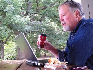 A photo of Tim Johnson, working on his computer, with the Arboretum in the background.