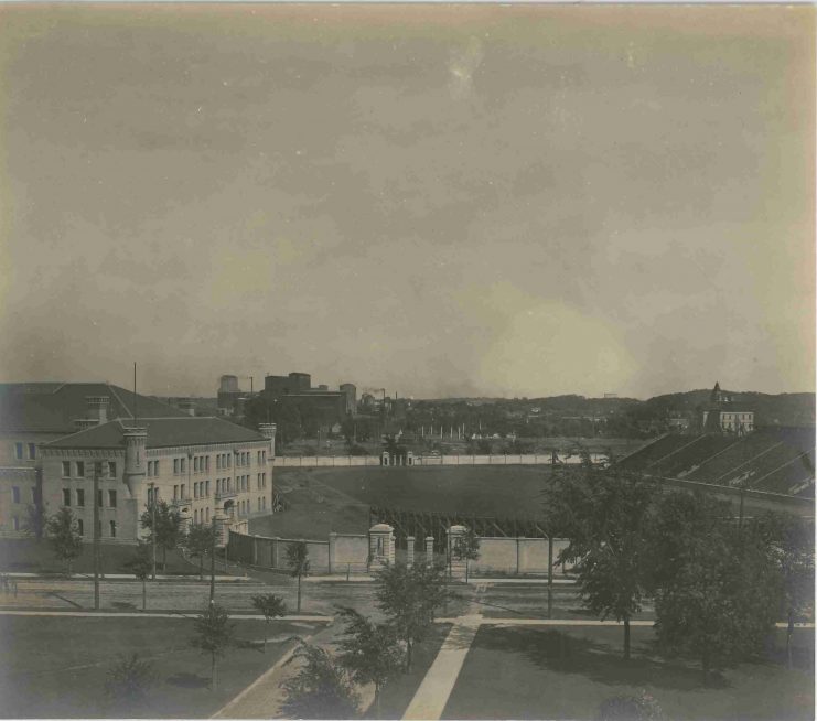Campus view showing Northrop Filed and its brick wall and entrance gates, circa 1906. The Armory is pictured to the left.