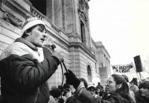 Black and white photo shows a crowd of student protestors holding signs to support the former General College. A protestor in the foreground speaks into a microphone to address the group.