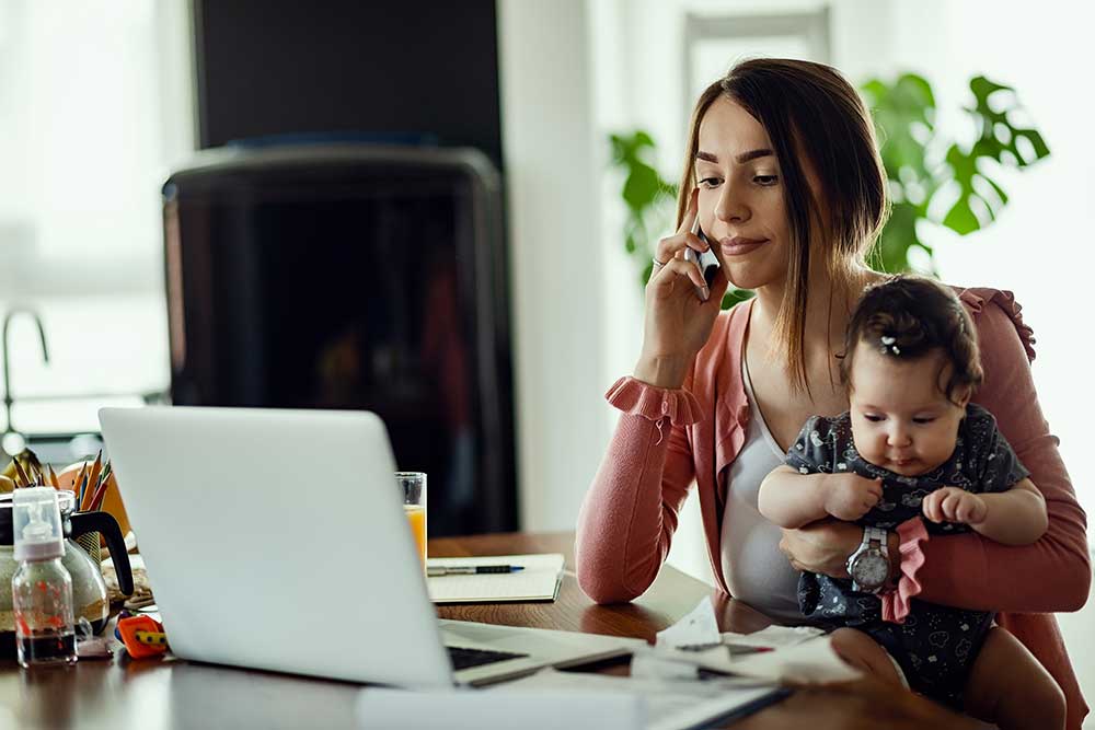 Woman on the phone and looking at her laptop while holding her baby girl on her lap.