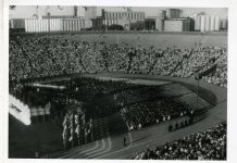 Graduates sit in chairs on the field of Memorial Stadium in front of a stage. Attendees are in the stadium bleachers.