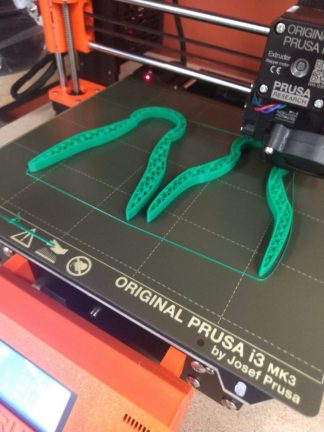 Two green tong devices are shown being printed by a 3D printer