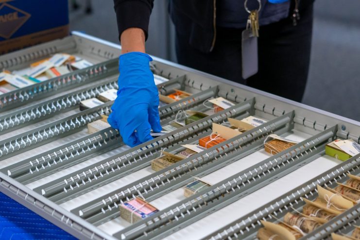 Anna Shepherd's blue-gloved hand is seen placing pharmaaceutical boxes in a large metal tray.