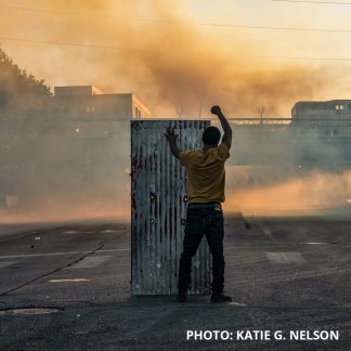 Photo by Katie G. Nelson of a person with raised fist with tear gas in the background