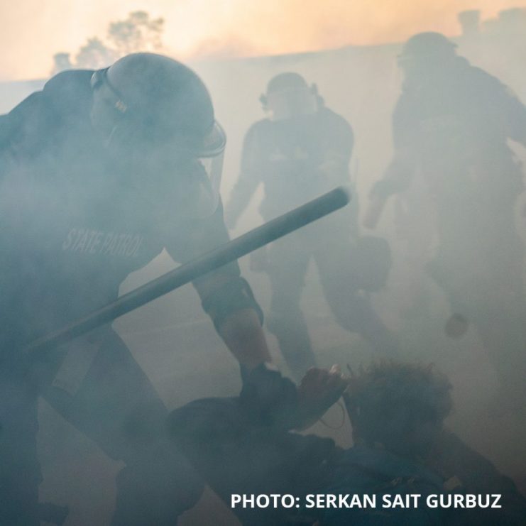 Photo by Serkan Sait Gurbuz of police in riot gear, gas mask, and wielding a metal bar, grabbing the wrist of a protestor who is kneeling down with hands behind their head