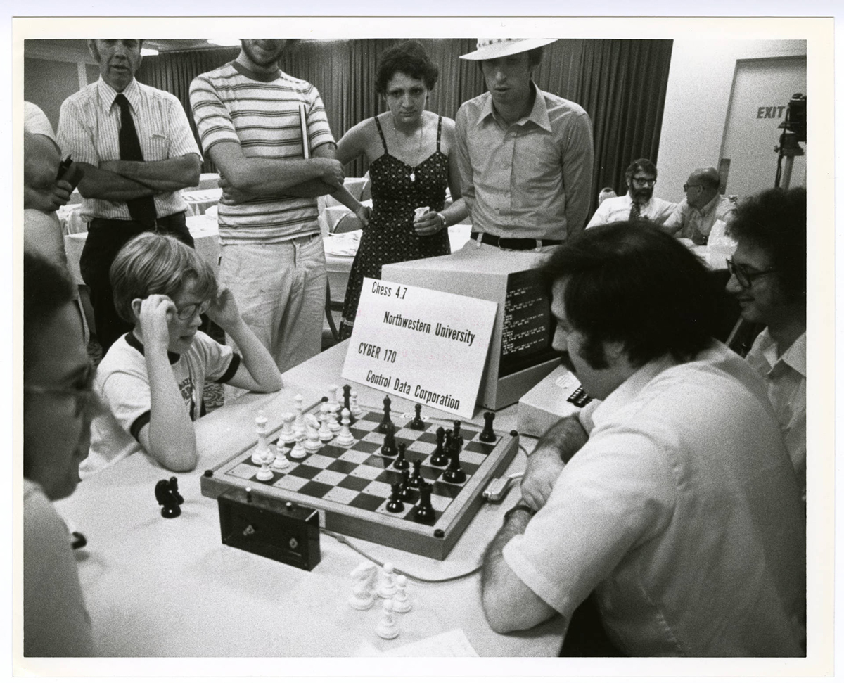 A black and white photo of a chess game in which two computer programs are competing with each other. The programs are Chess 4.7 from Northwestern University and Cyber 170 from Control Data Corporation. People are seated on either side of the chessboard to move the chess pieces.