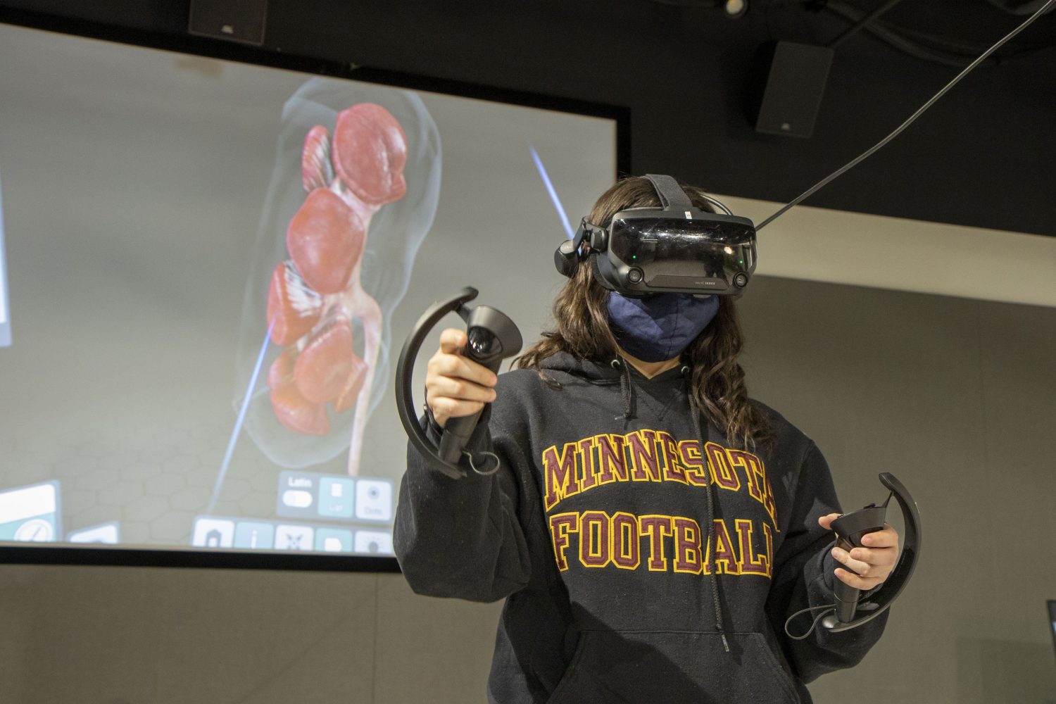 A person wearing a virtual reality headset stands in front of a screen showing an anatomical representation of an organ