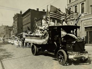A photo from the Social Welfare History Archives showing a parade on a cobblestone street. In the foreground, an early 20th-century motor vehicle has a flatbed in which many people are riding while holding American flags