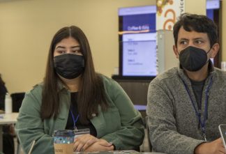 Two people, masked, at a conference, with computer screen behind.