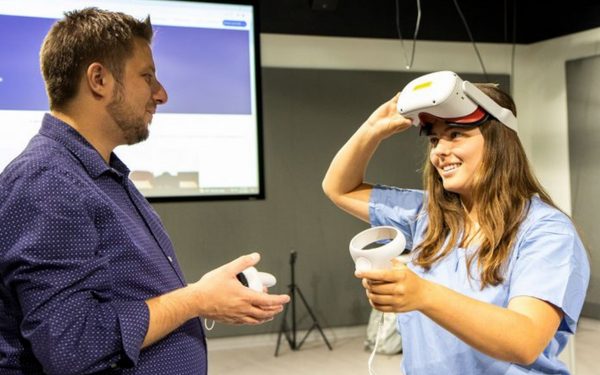 Two people facing each other. One person (right) has a virtual reality headset on their head and a controller in their hand. The other person (left) has a virtual reality controller in their hand.
