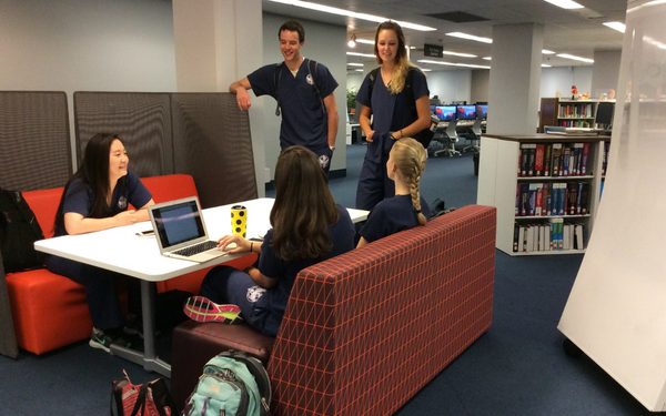 Five students wearing scrubs sitting and standing around a table with benches in a library.