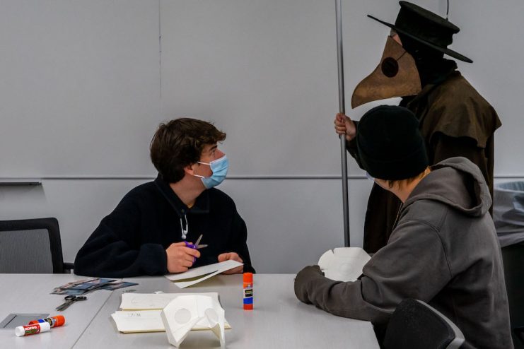 Two people are seated at a table and using scissors to cut out Plague doctor masks while the Plague Doctor watches their progress.