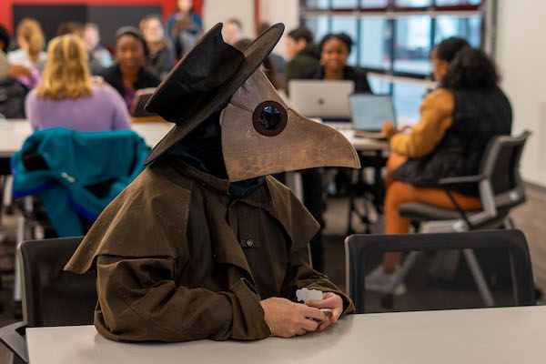 A person wearing a Plague Doctor costume (black hat, mask with large eye holes and a bird's beak, brown trench coat) sits at a classroom table during a lecture.