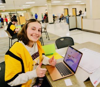 Young woman giving two thumbs up, in library setting with laptop.