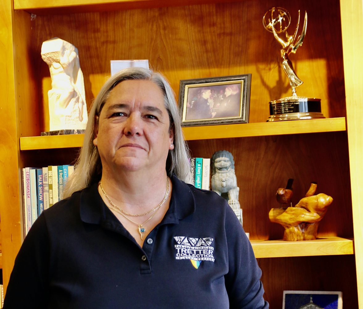 Woman wearing blue polo t-shirt bearing the logo "Tretter Collection" standing in front of a bookcase, where several book spines and a gold Emmy statue are visible